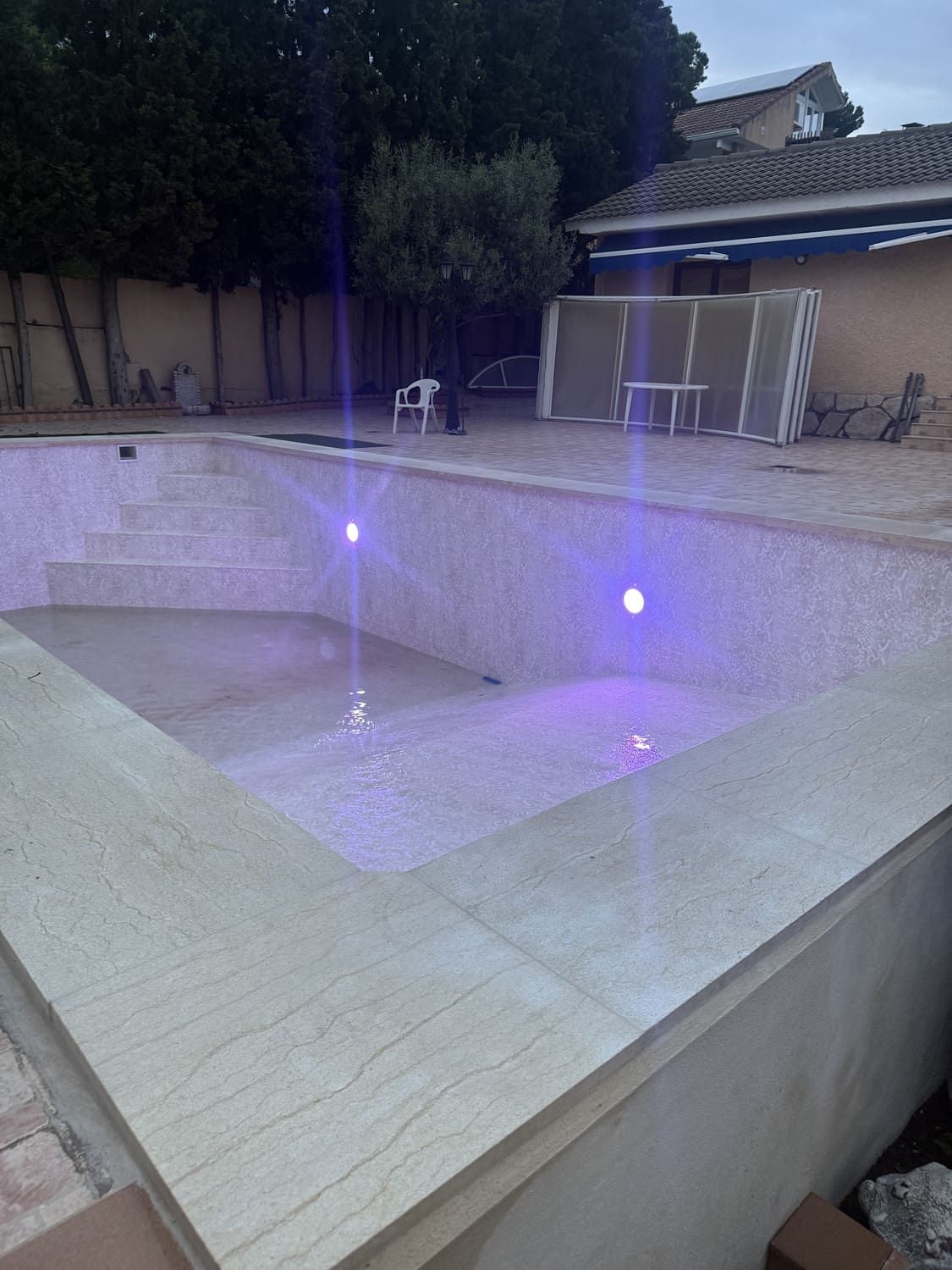 Piscina con iluminación violeta, borde de piedra e interior de azulejos. Escenario nocturno con árboles al fondo.