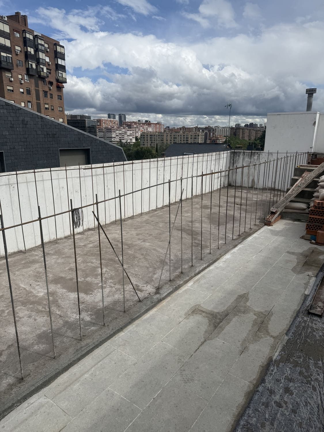 Azotea en construcción con piso de concreto, varillas de metal y vista a edificios y cielo.
