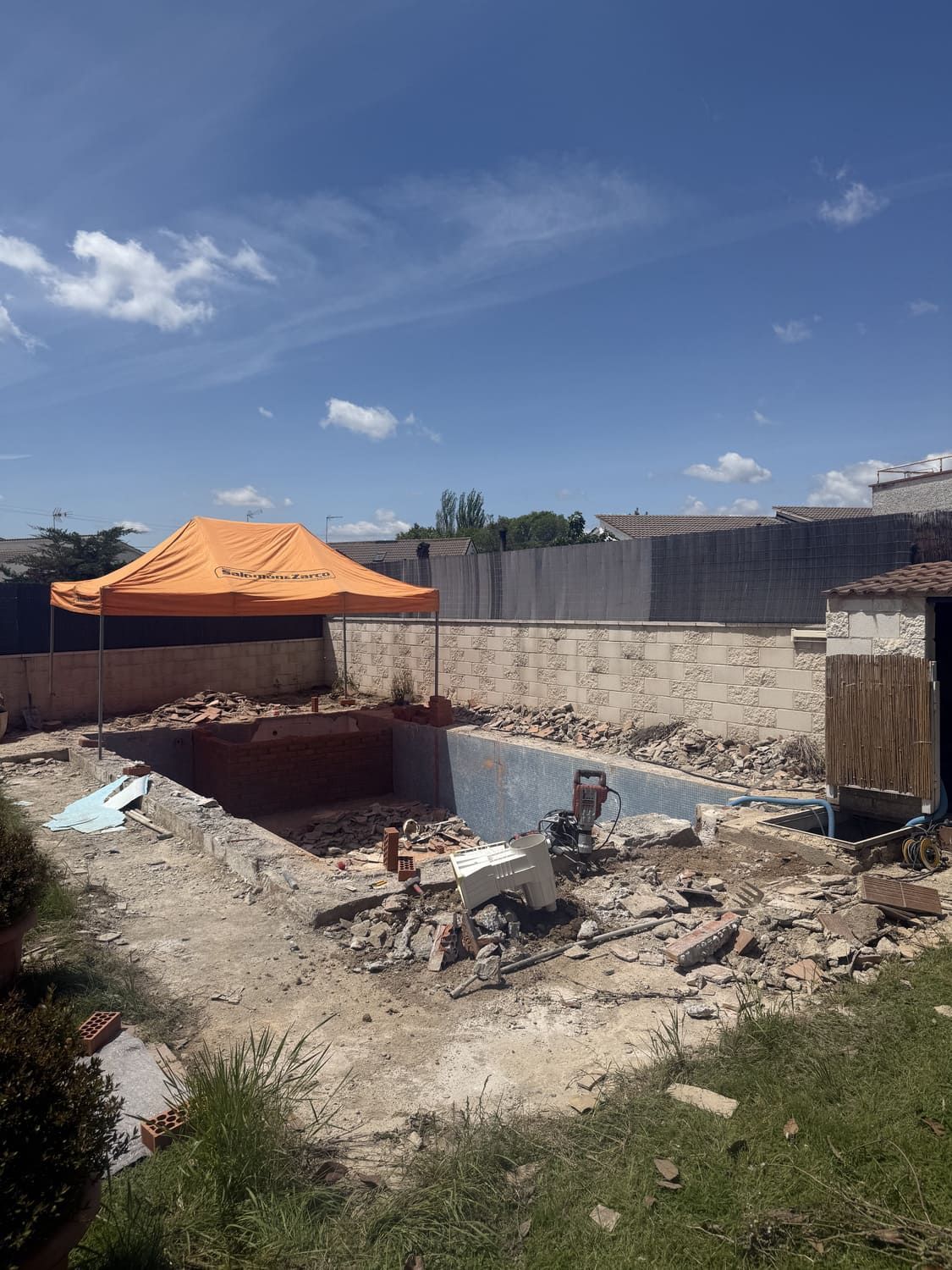 Demolición de piscina en progreso bajo un cielo azul; escombros esparcidos, carpa naranja en el fondo.