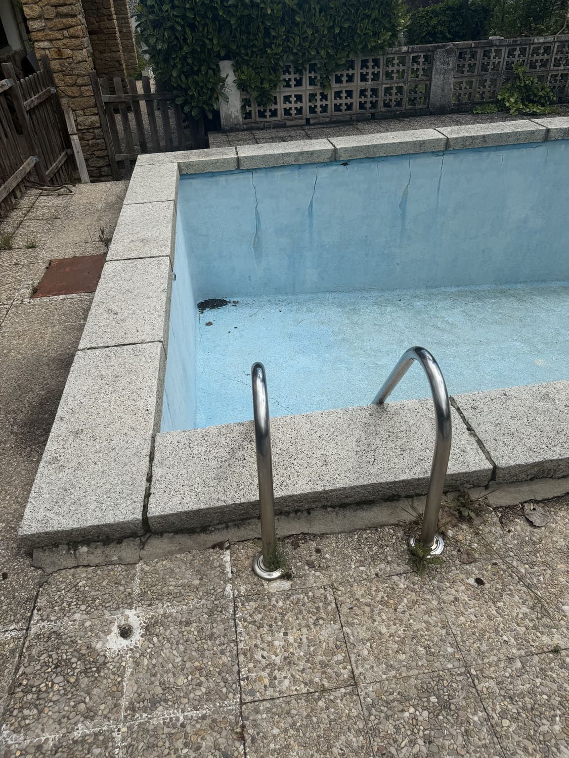 Piscina exterior rectangular, vacía, con interior azul y escalera metálica. Borde de piedra, patio de hormigón y vegetación al fondo.