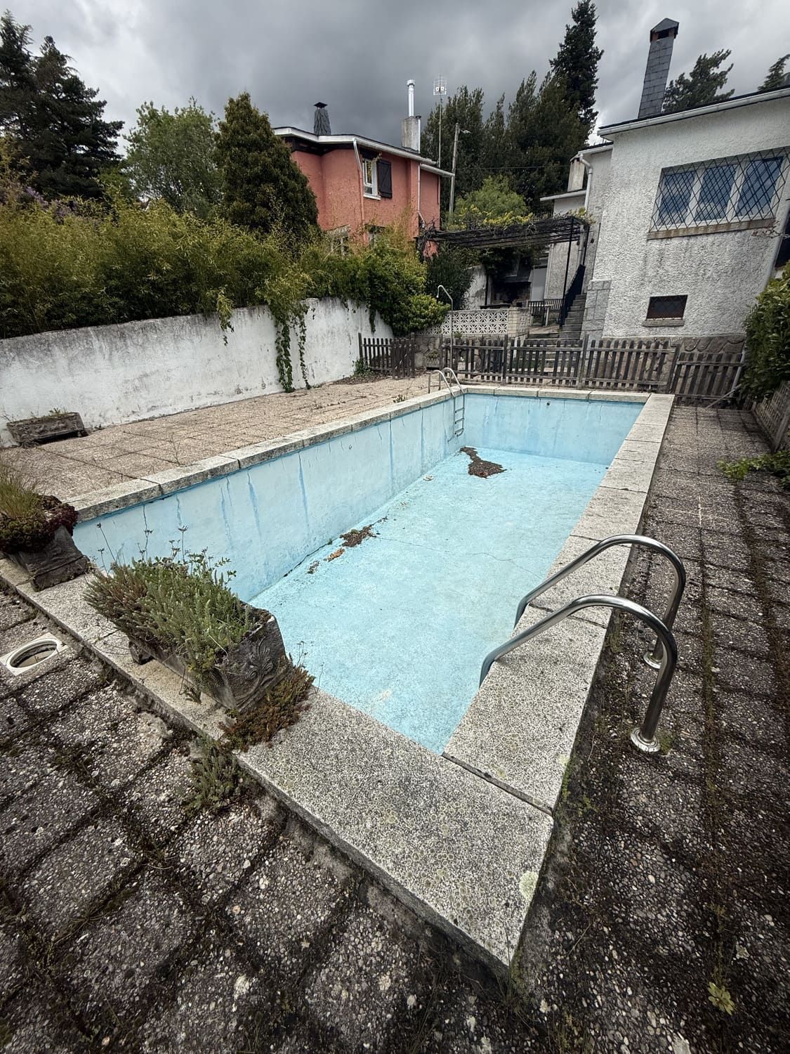 Una piscina abandonada, con agua sucia, rodeada de piedras antiguas y plantas crecidas.