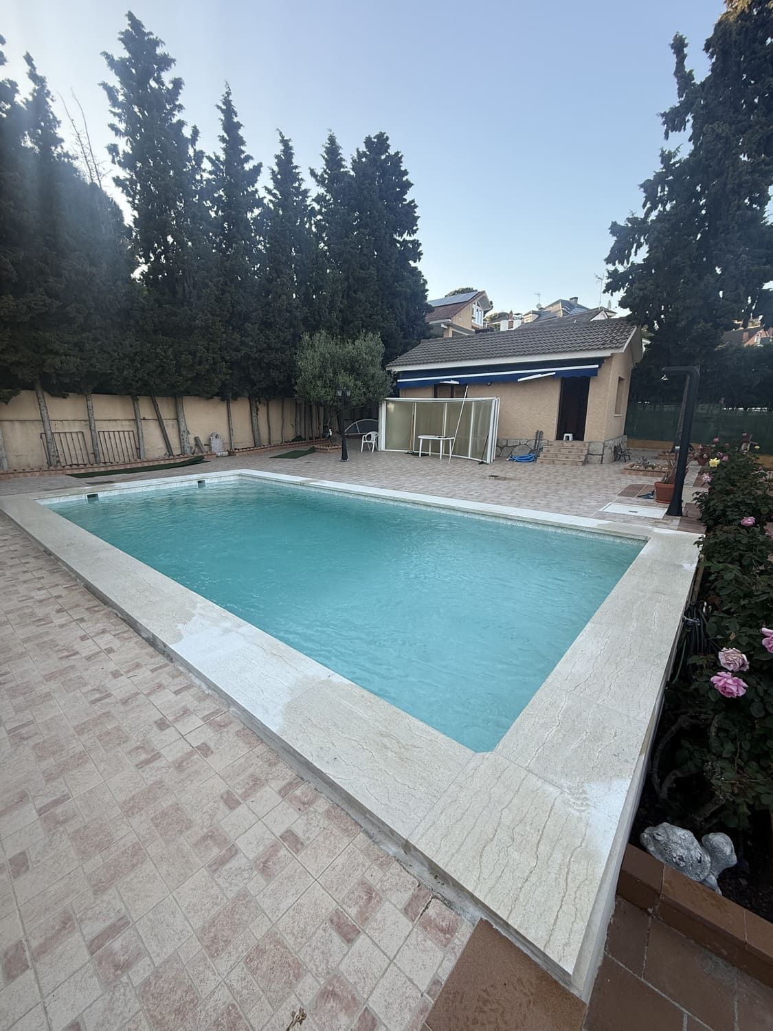 Una piscina rectangular de agua turquesa, rodeada de adoquines de color claro. Edificios y árboles al fondo.