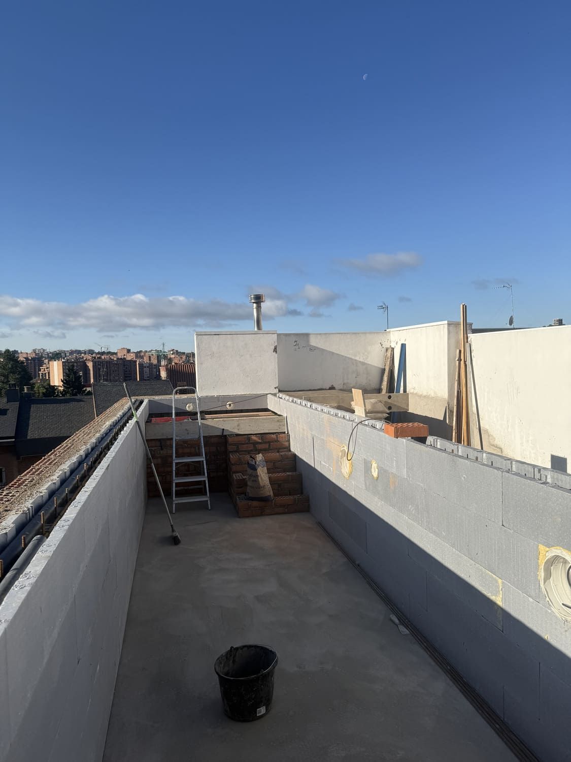 Una azotea con paredes blancas y un cielo azul despejado. Se ven una escalera, ladrillos y un cubo negro.