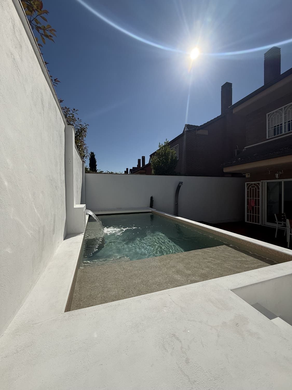 Pequeña piscina rectangular con fuente, rodeada de paredes blancas en un día soleado.