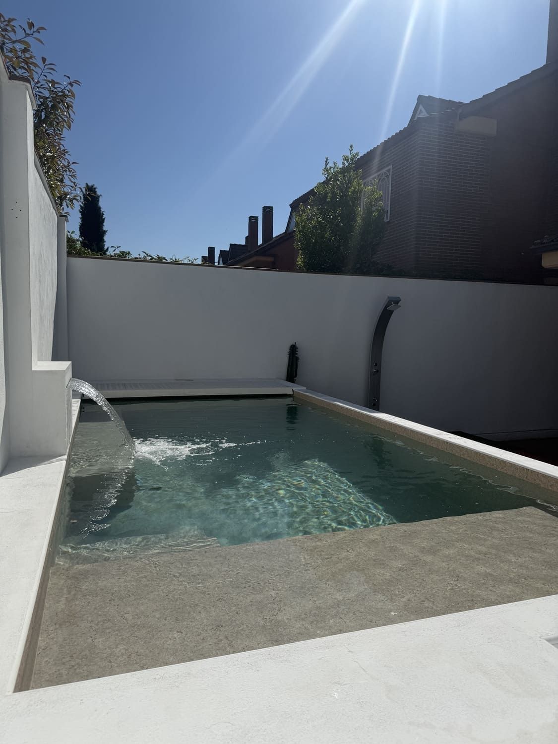 Pequeña piscina rectangular con cascada de agua, rodeada de paredes blancas bajo un cielo azul brillante.