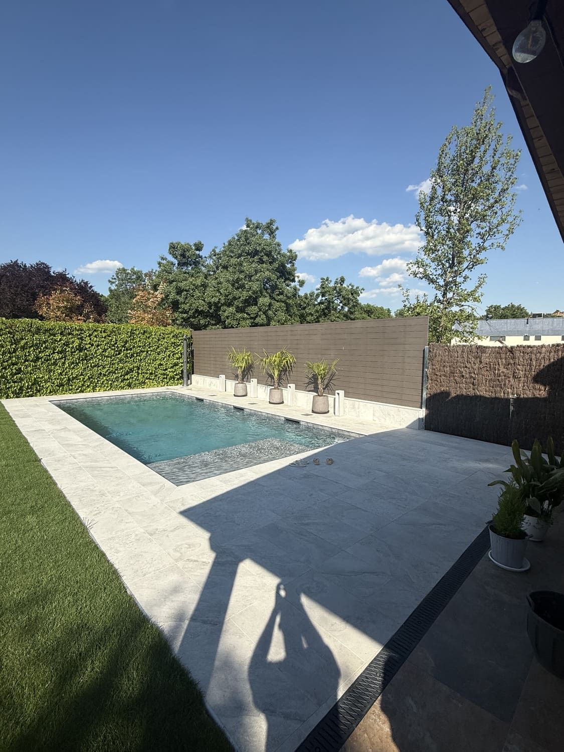 Patio trasero con piscina, terraza, vegetación y cielo azul. La sombra de una persona tomando la foto.