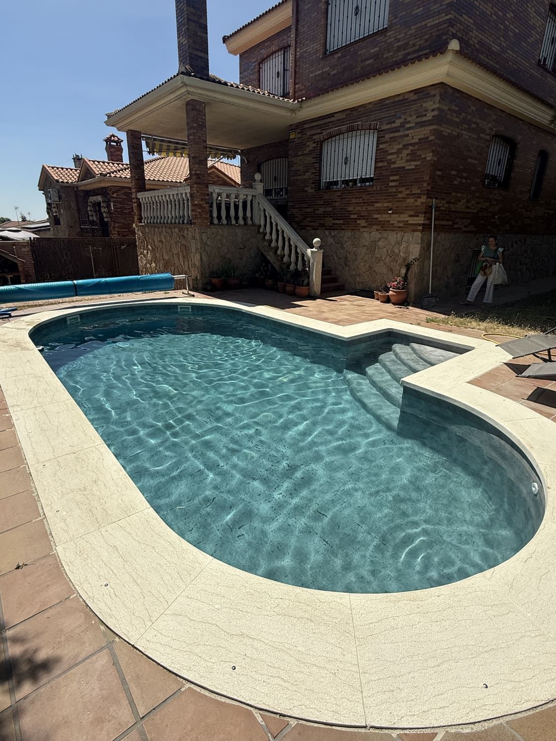 Piscina frente a una casa de ladrillo con escaleras y porche cubierto. Día soleado.