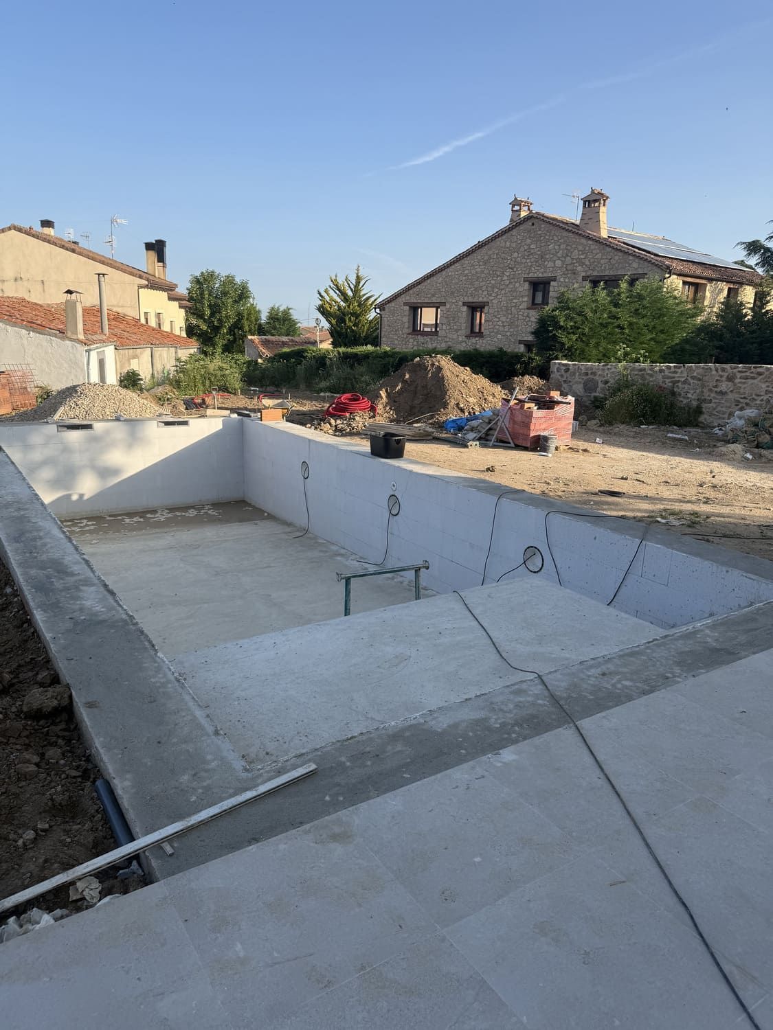 Piscina de hormigón en construcción, con escalones y materiales de construcción a su alrededor. Casas al fondo bajo un cielo azul.