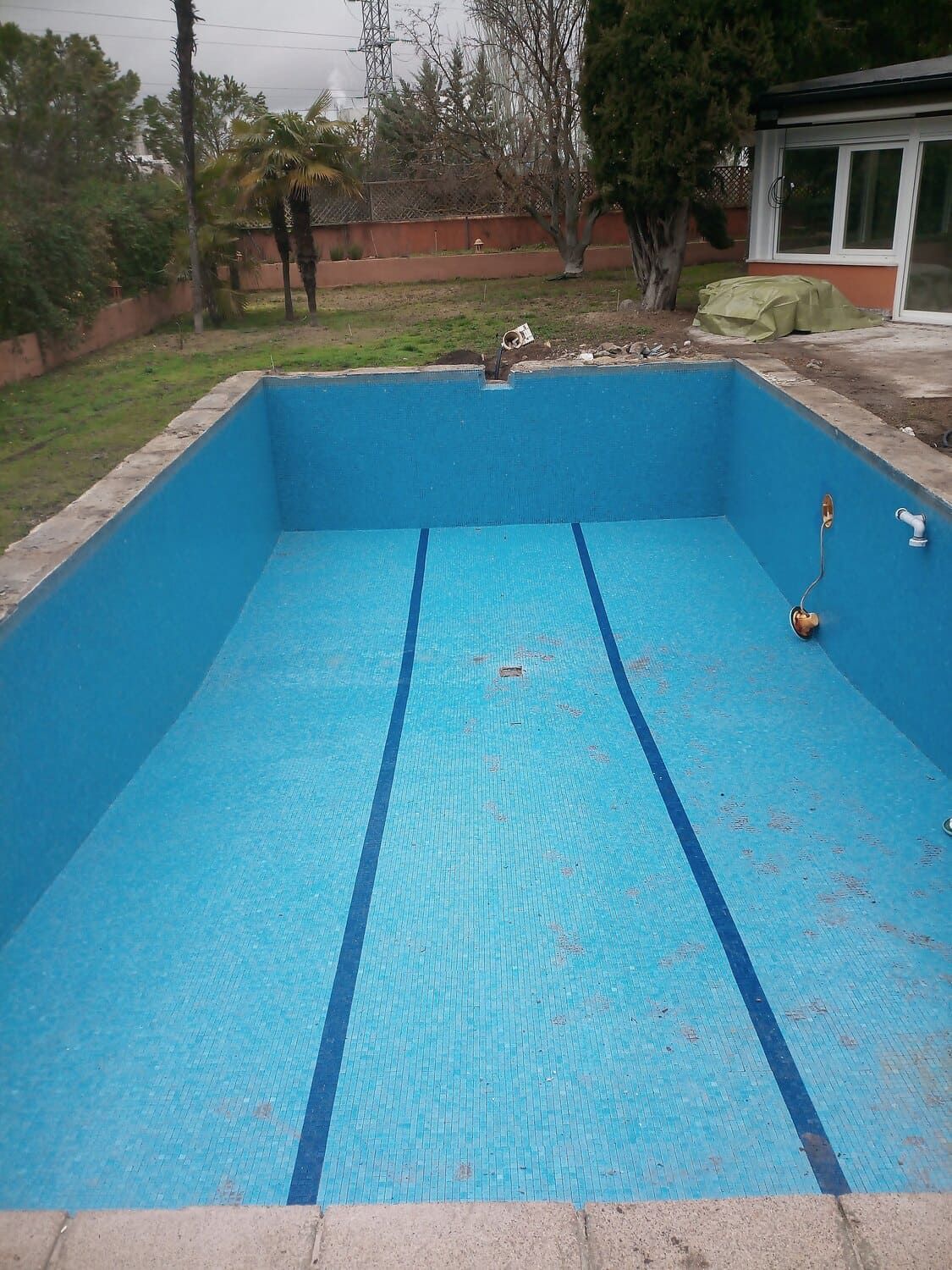 Piscina vacía de azulejos azules en un patio trasero, con marcadores de carril y hormigón circundante.