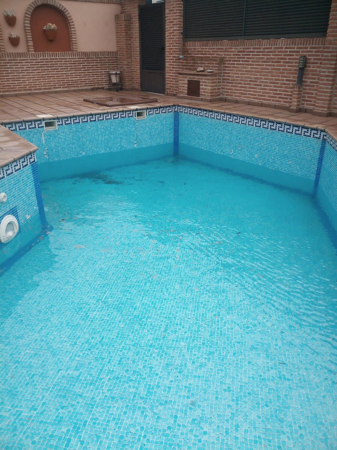 Una piscina de azulejos azul claro, casi vacía, al aire libre. Paredes de ladrillo marrón la rodean.