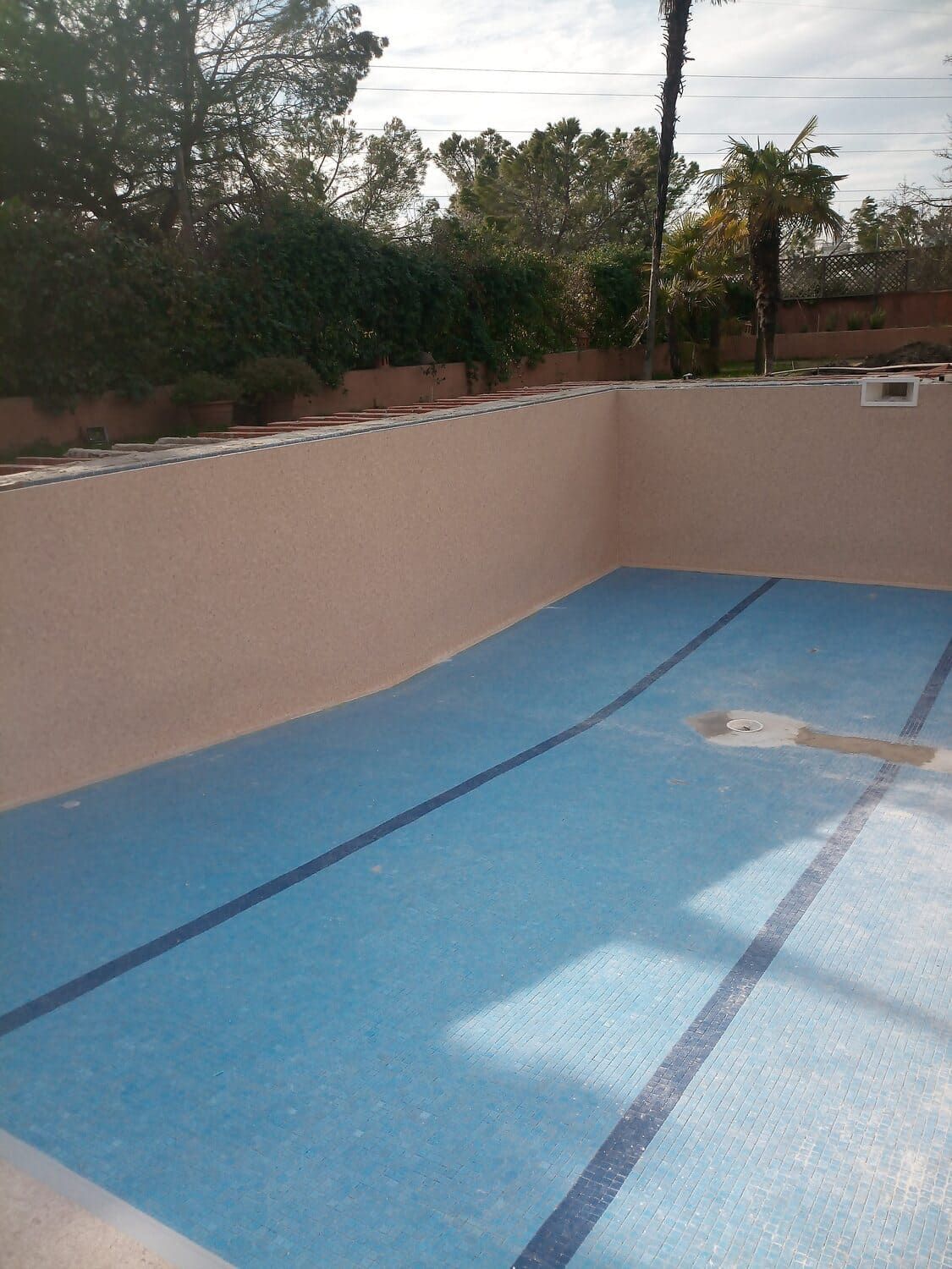 Piscina vacía con fondo de azulejos azules, paredes beige y árboles alrededor.