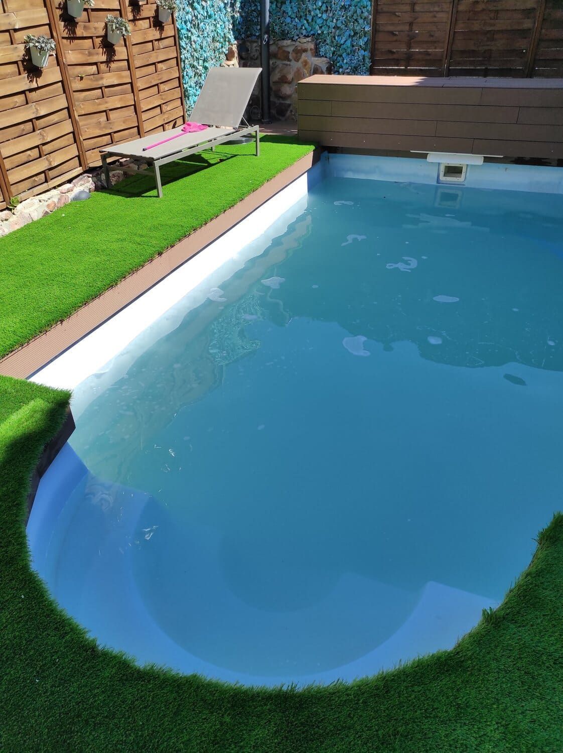 Una piscina rectangular de agua azul claro, rodeada de césped artificial y un deck de madera.