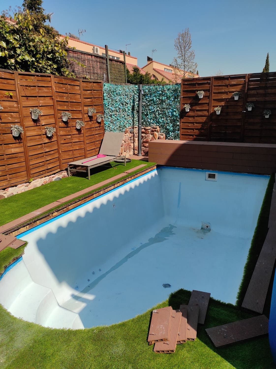 Piscina vacía con líneas azules en un patio trasero con vallas de madera, césped artificial y tumbonas.