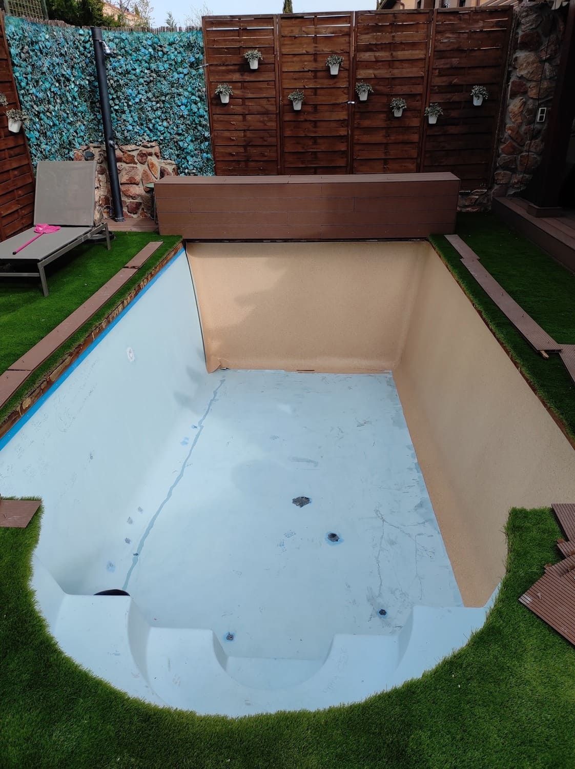 Piscina rectangular vacía, con interior azul claro, rodeada de césped y baldosas marrones. Valla de madera al fondo.