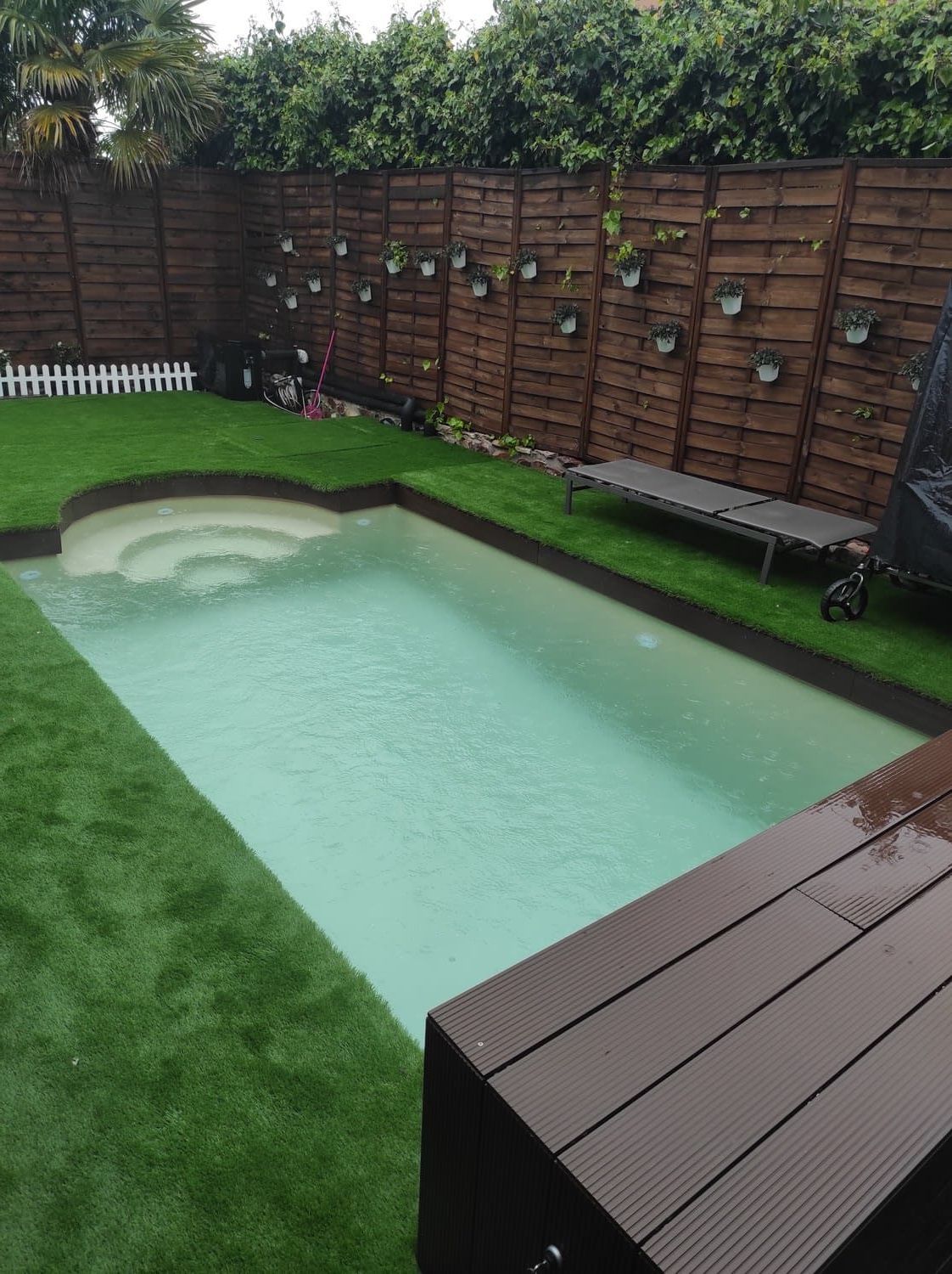Una pequeña piscina en el patio trasero con escalones y césped verde circundante, terraza y cerca de madera marrón y plantas colgantes.