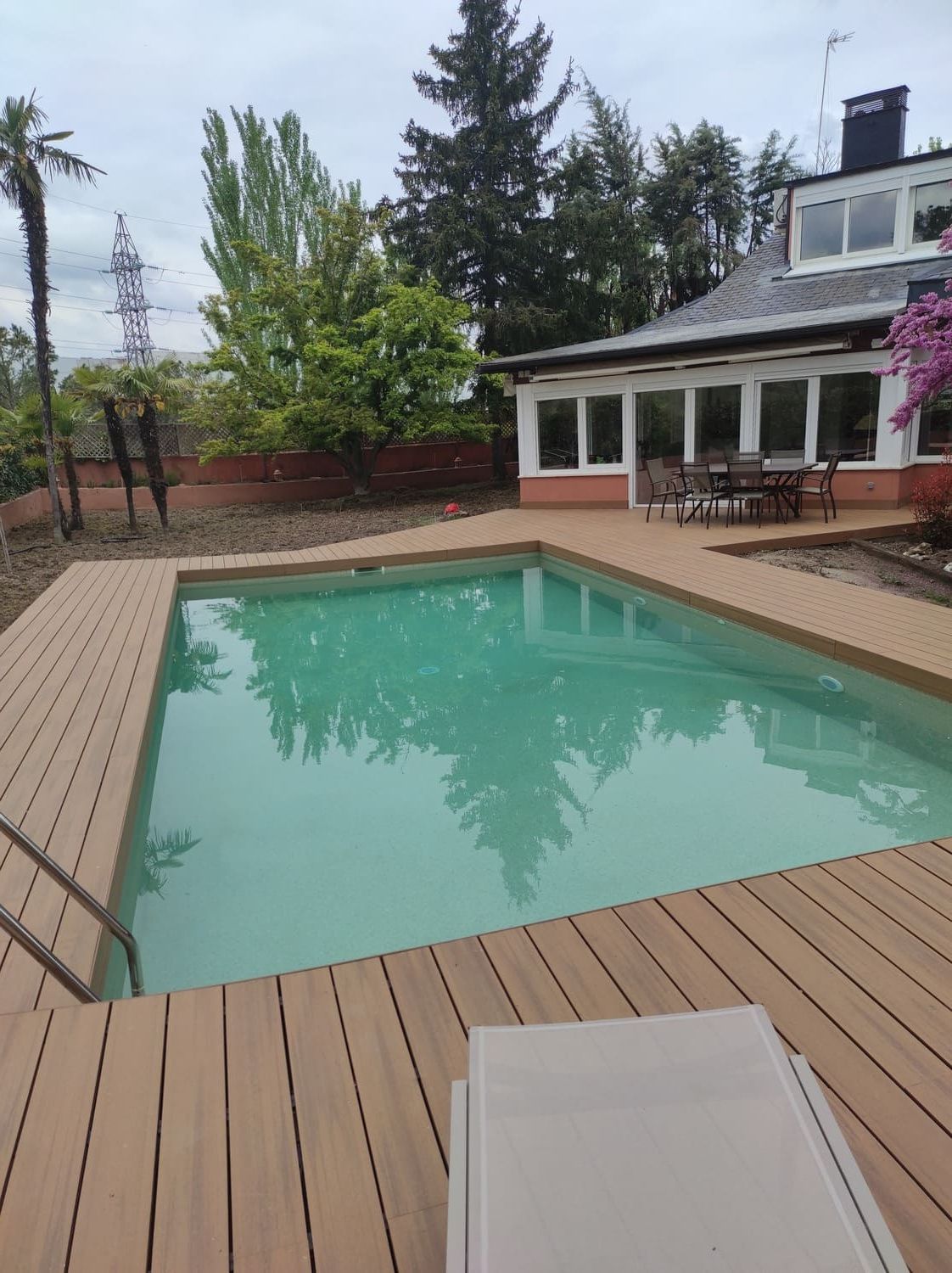 Piscina rodeada de tarima de color marrón claro, frente a una casa blanca con grandes ventanales.