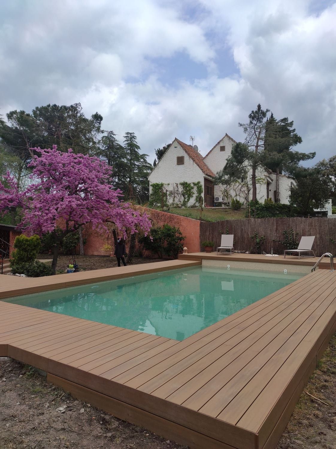 Piscina con terraza de madera, casa blanca al fondo, árbol en flor rosa, cielo nublado.