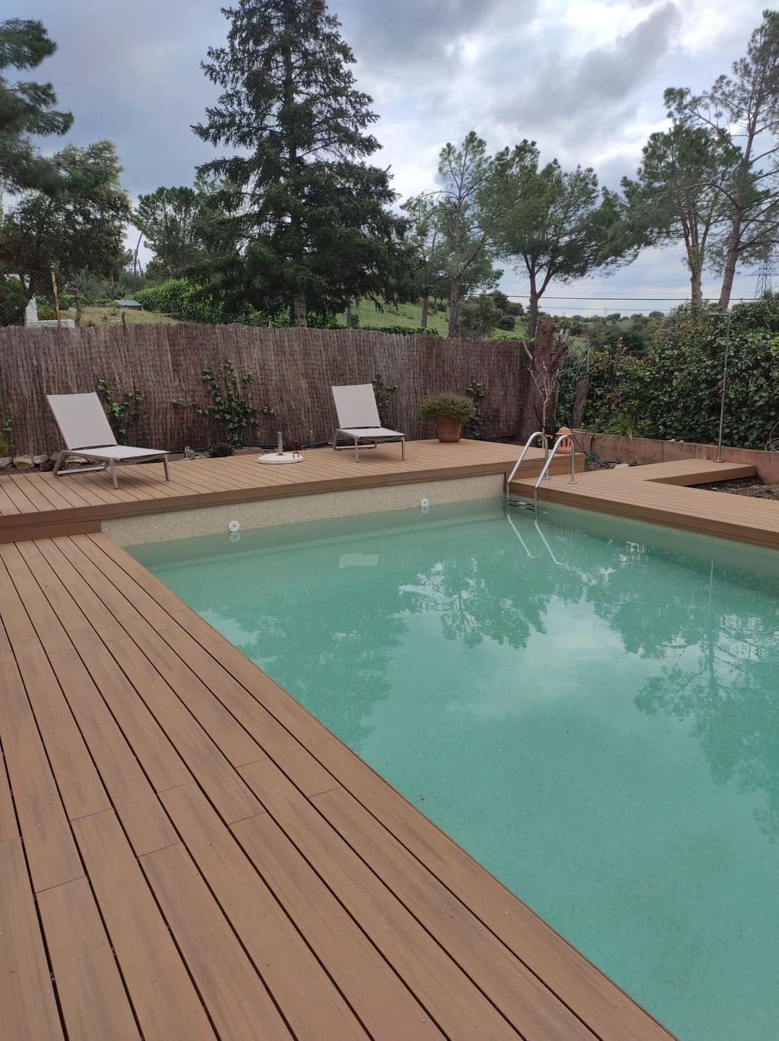 Piscina con deck de madera y camastros, rodeada de vegetación y un cielo nublado.
