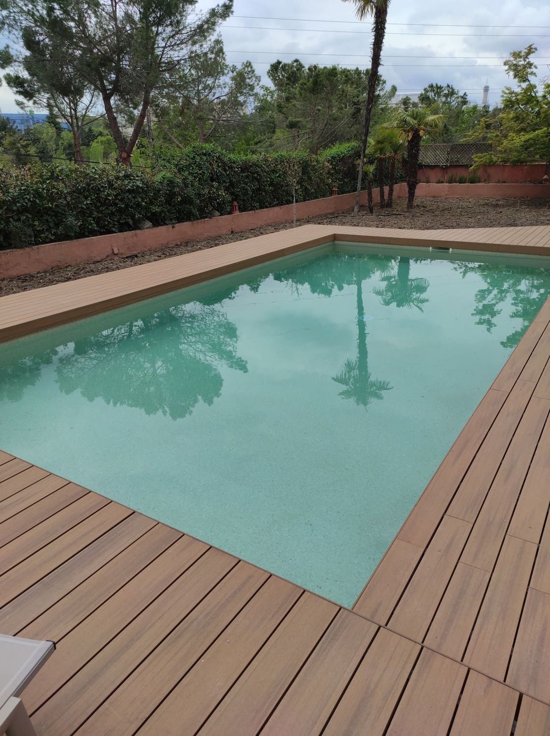 Piscina rectangular rodeada de tarima de madera. Palmeras reflejadas en el agua. Exuberante vegetación al fondo.