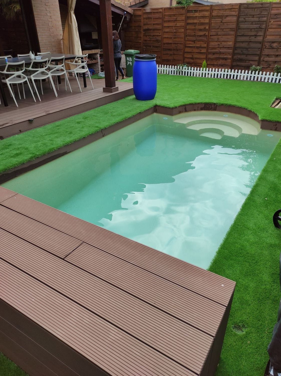 Una pequeña piscina rectangular con escalones de color marrón rodeada de césped artificial y un deck de madera.