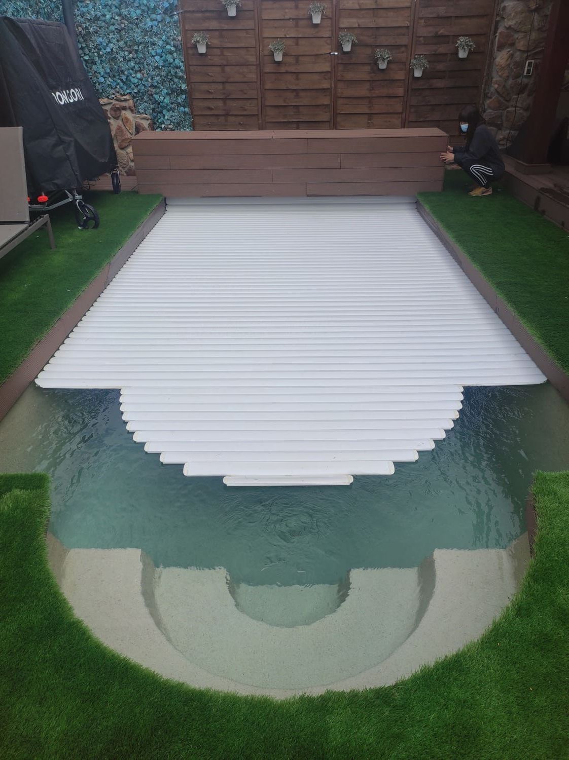 Piscina con cubierta retráctil blanca, rodeada de césped artificial. Al fondo, una valla de madera.