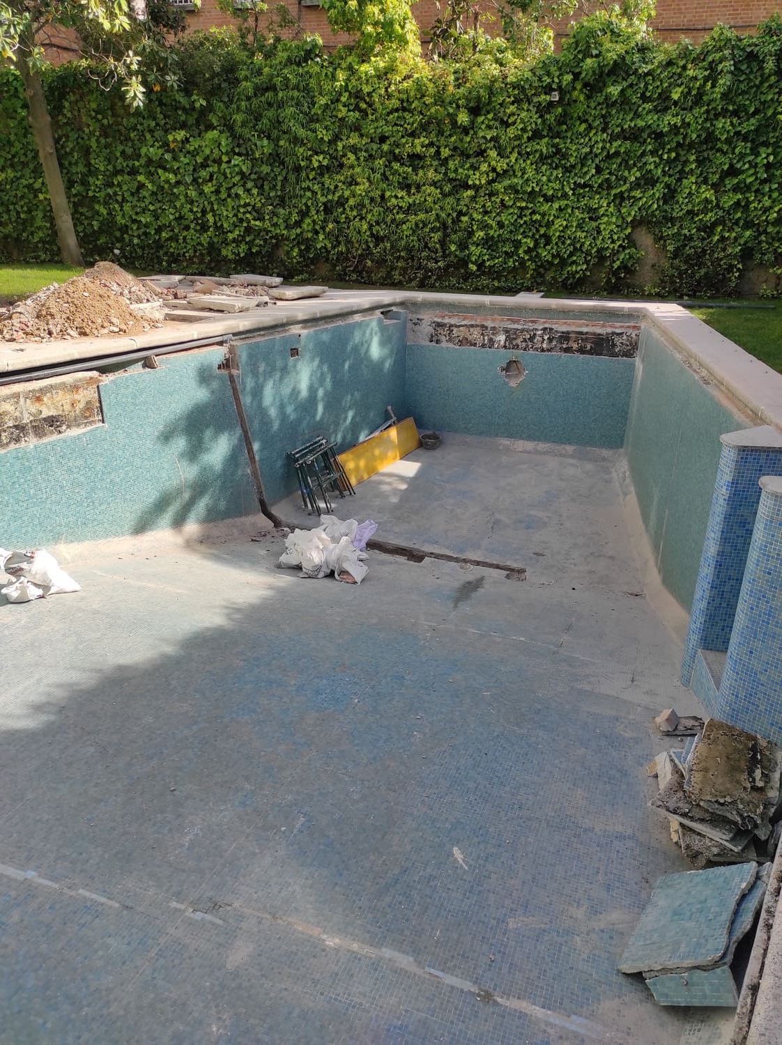 Piscina en construcción, con azulejos y hormigón a la vista. Hay herramientas y escombros. Un seto verde y frondoso al fondo.