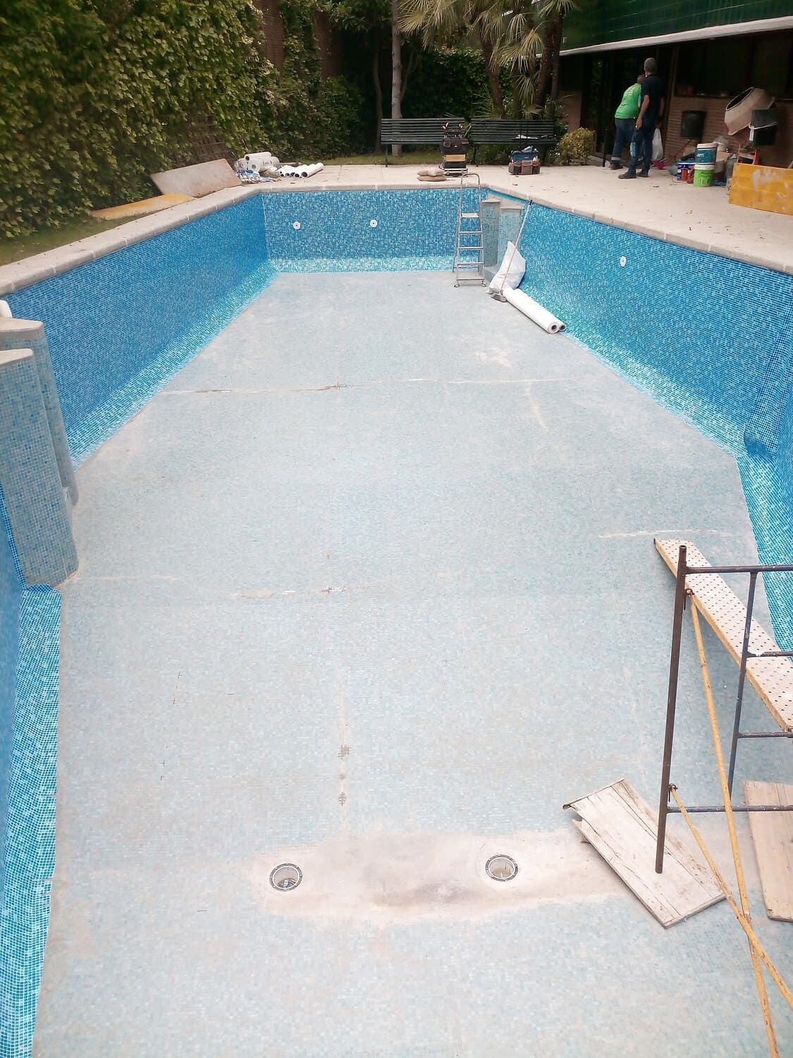 Piscina rectangular vacía en construcción con paredes de azulejos azules y suelo gris claro; trabajadores de la construcción en el fondo.