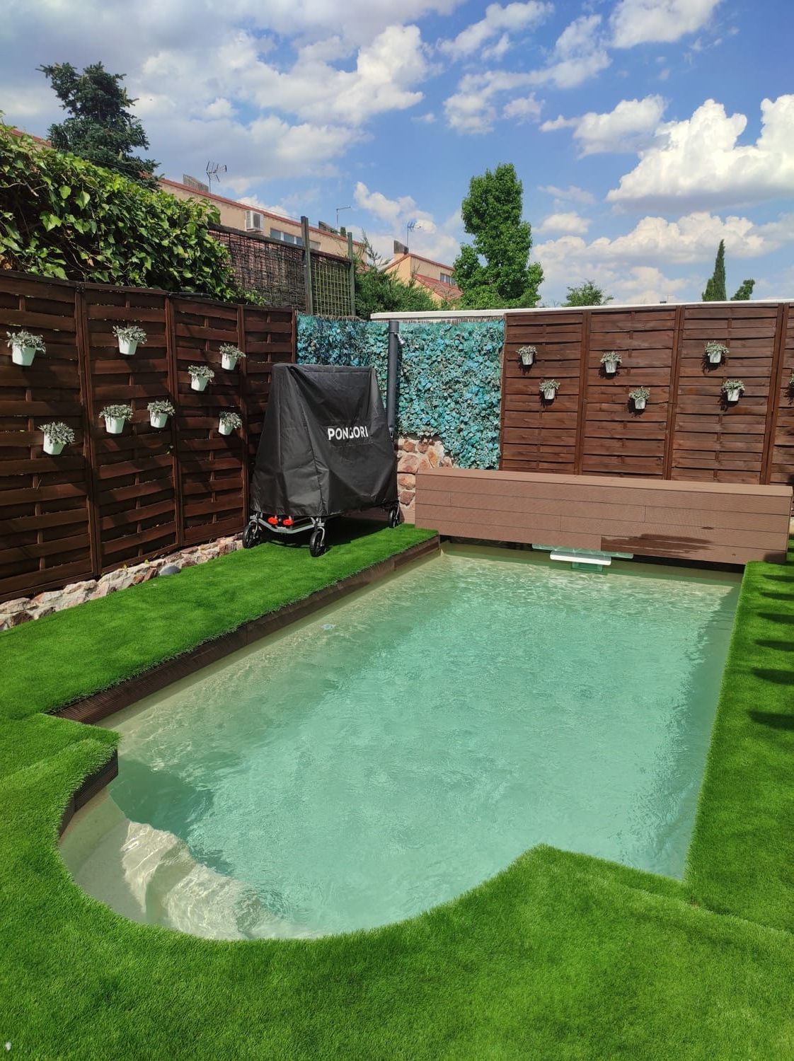 Pequeña piscina turquesa en un patio trasero con césped artificial verde. Cerca de madera marrón con jardineras colgantes y una parrilla cubierta.