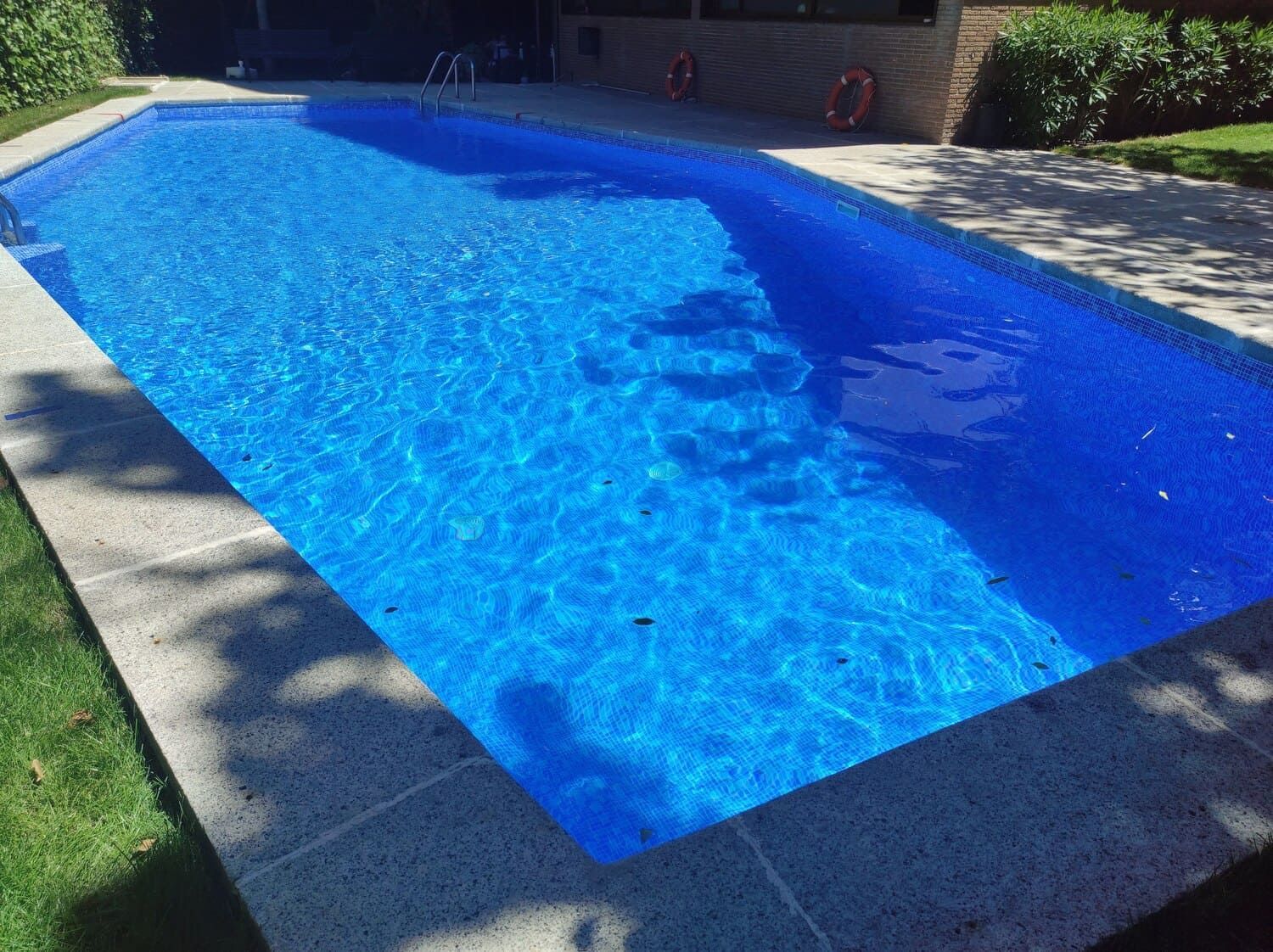 Piscina rectangular llena de agua azul brillante, rodeada de piedra gris y césped verde.