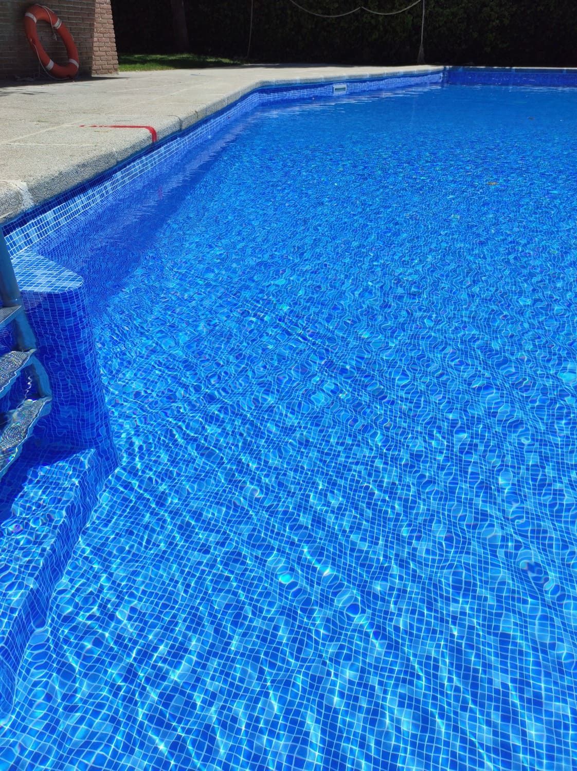Piscina de azulejos azules con agua cristalina, escalones y un pequeño salvavidas rojo. Al aire libre.