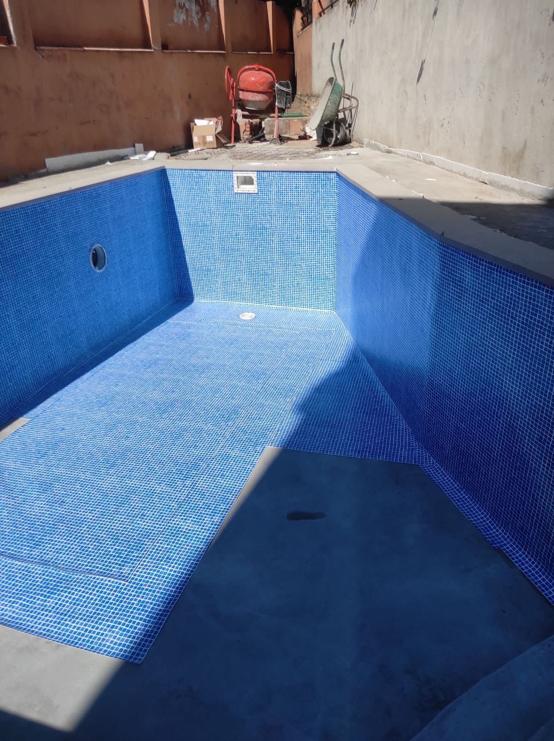Piscina vacía revestida con mosaicos azules. Herramientas y materiales de construcción visibles en el exterior.