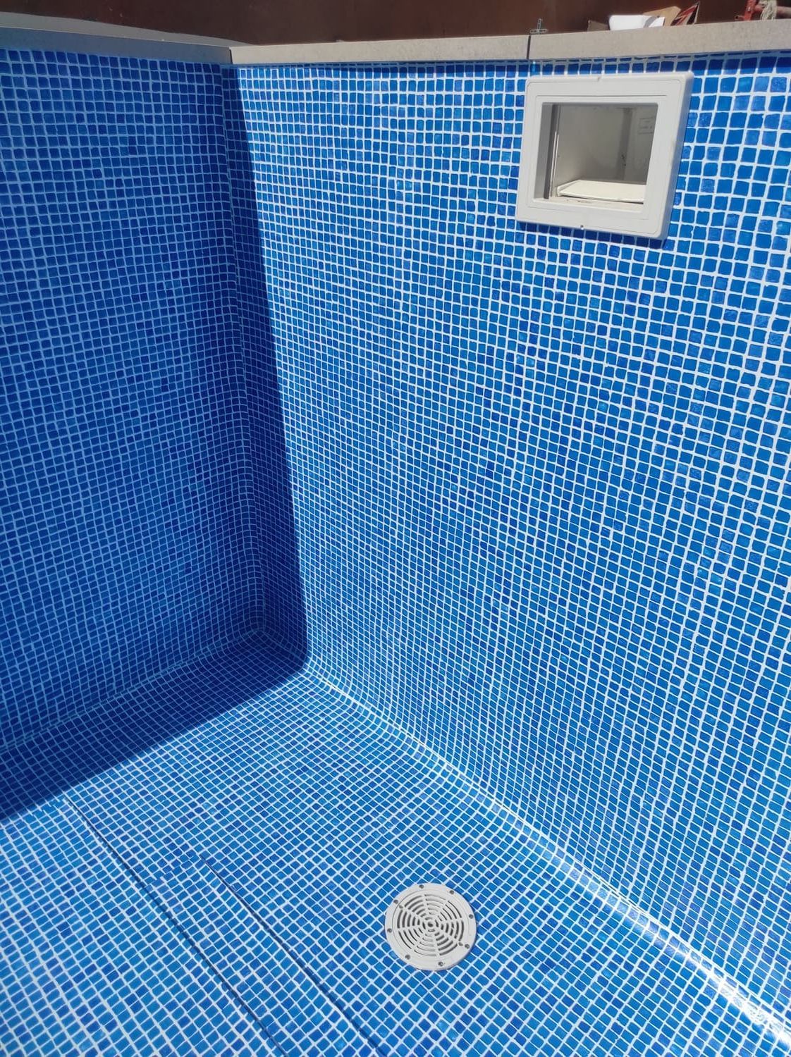 Interior de piscina con azulejos azules, desagüe, luz y luminaria rectangular de color blanco.