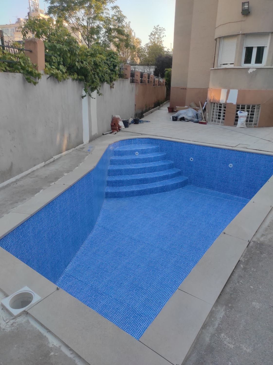 Piscina de azulejos azules con escalones, rodeada de hormigón y un edificio, al aire libre.