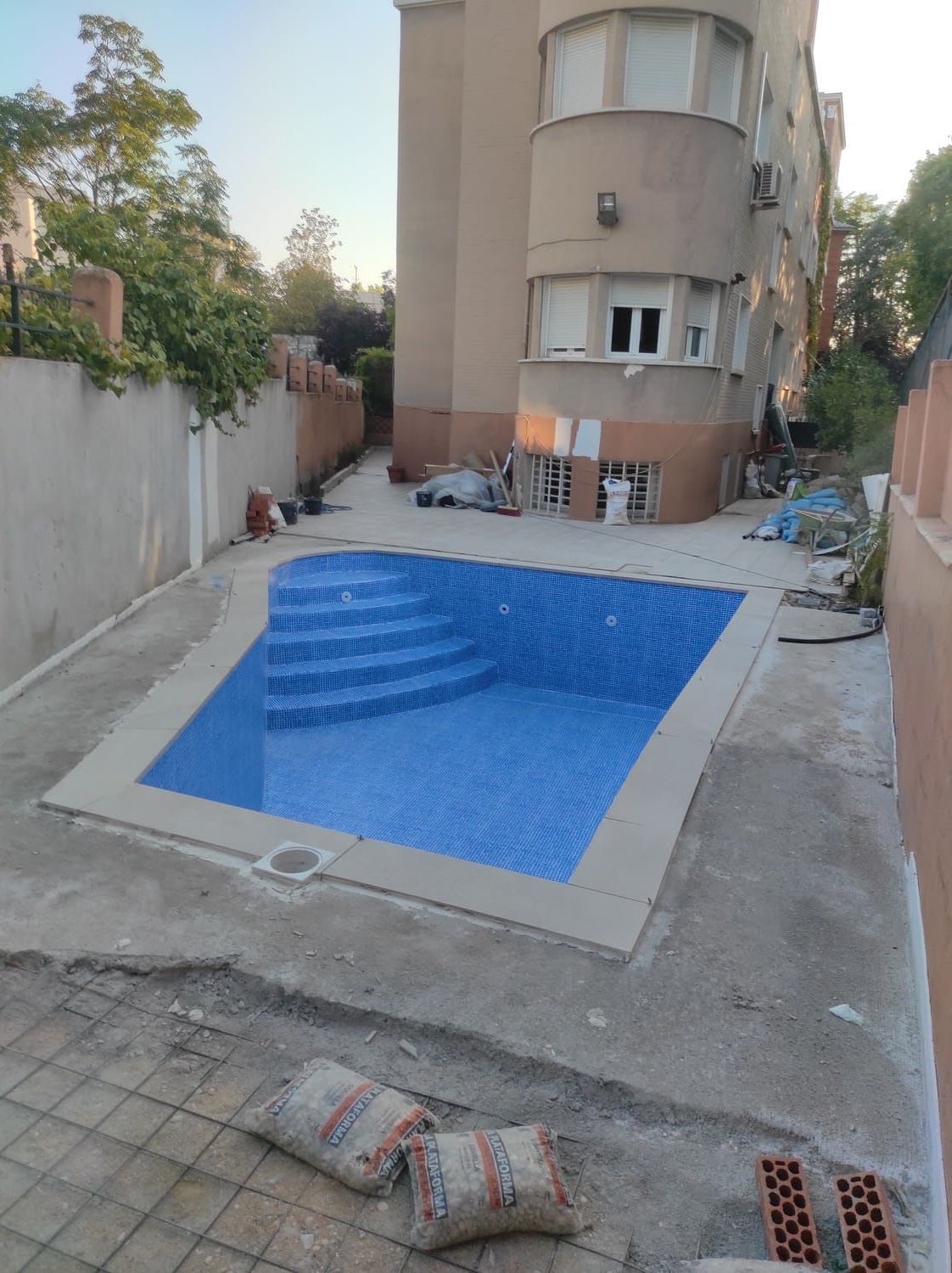 Una piscina a medio construir con azulejos azules, frente a un edificio. Dos sacos de cemento en el suelo.