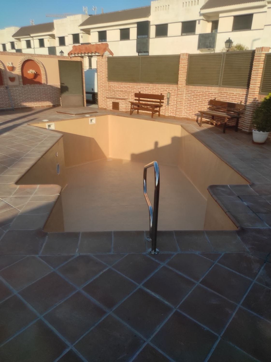 Piscina vacía de color beige rodeada de un patio de baldosas marrones y muros bajos de ladrillo. Dos bancos de madera se encuentran cerca.