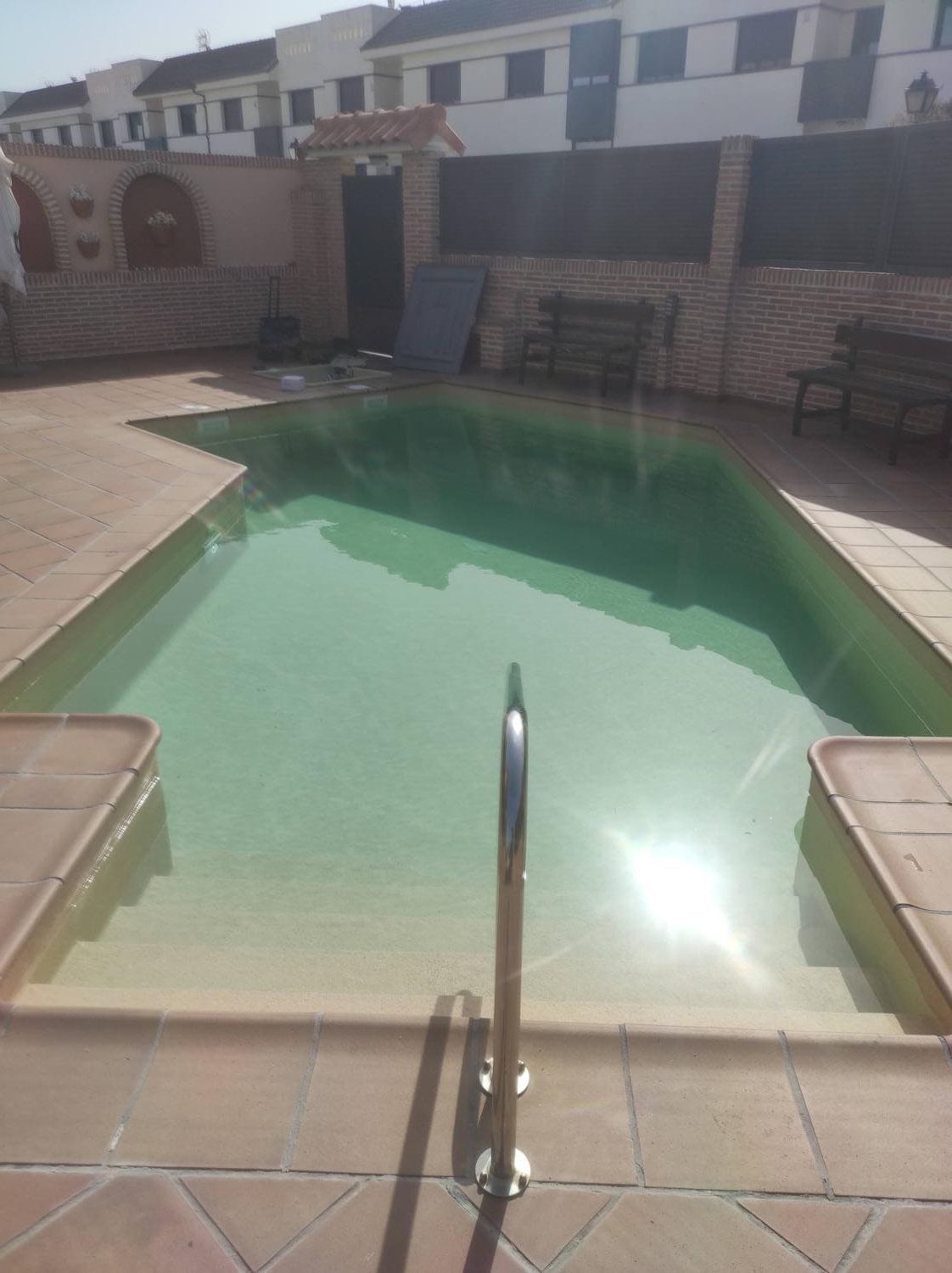 Piscina verde rodeada de baldosas color canela y escalones, con barandilla metálica y edificios al fondo.