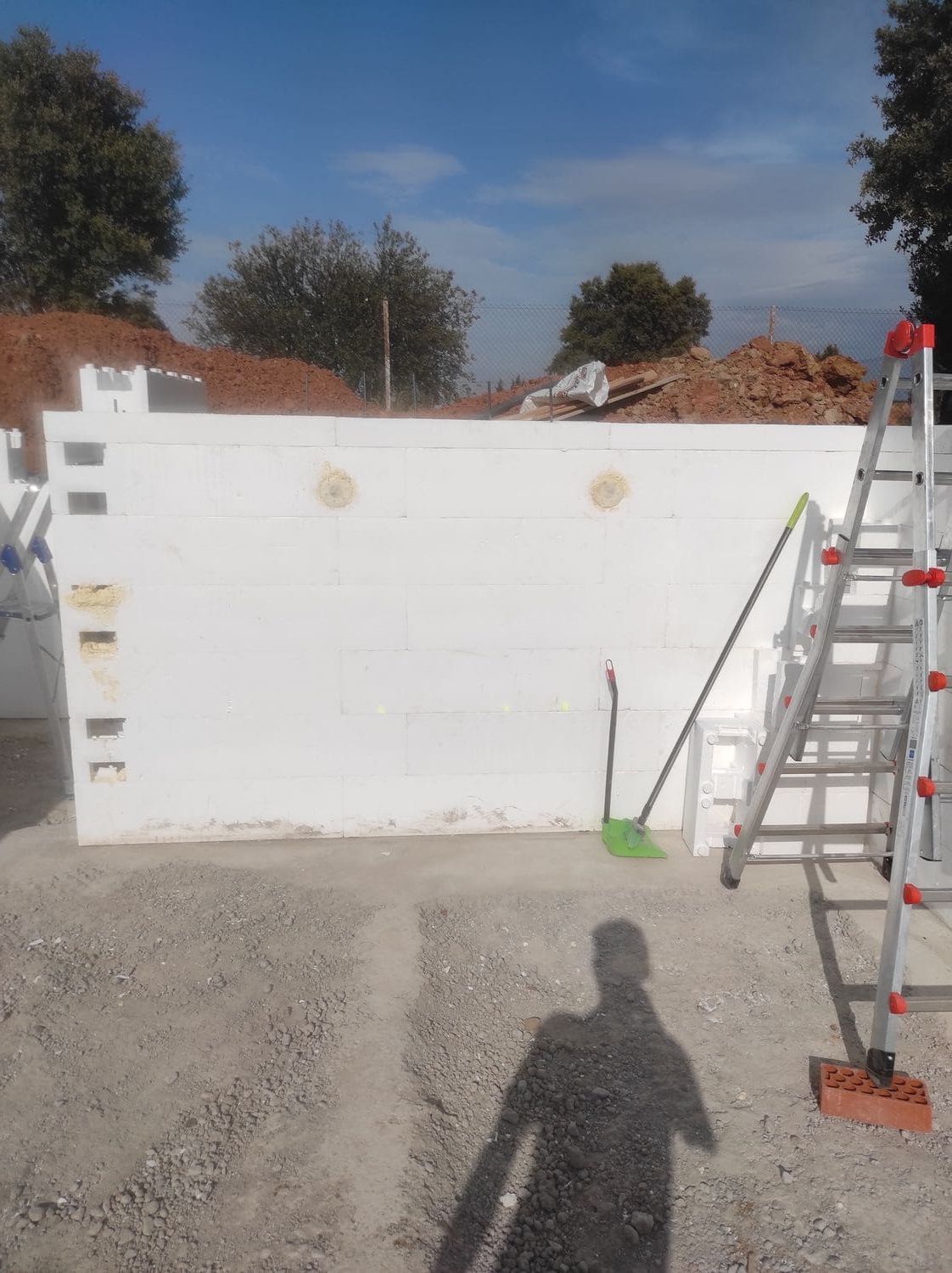 Muro de bloques de espuma blanca, herramientas apoyadas, escalera y la sombra de una persona. Exterior.