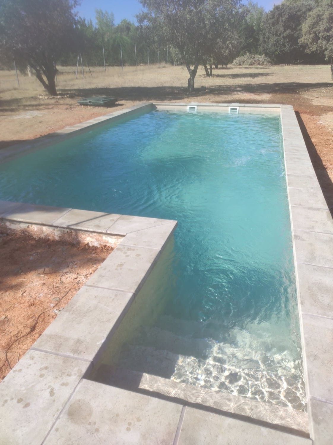 Piscina rectangular de color turquesa con escalones, rodeada de piedra y tierra, bajo un cielo soleado.