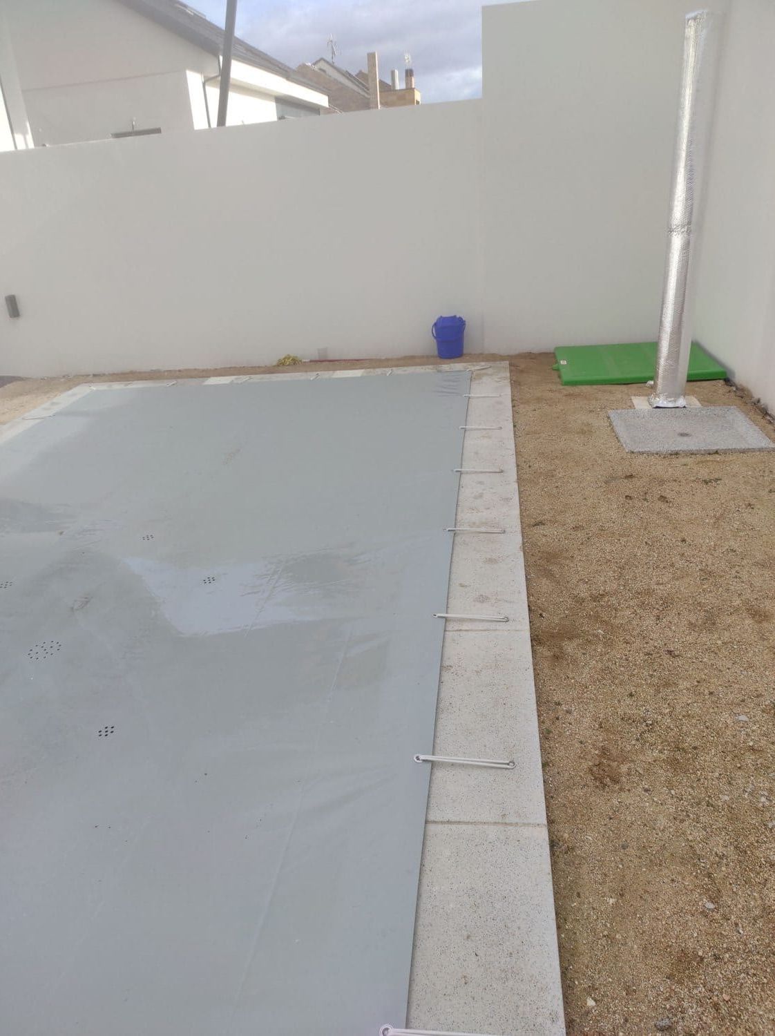 Una piscina rectangular cubierta con una lona gris se encuentra junto a una zona de grava. Al fondo se ve una pared blanca.