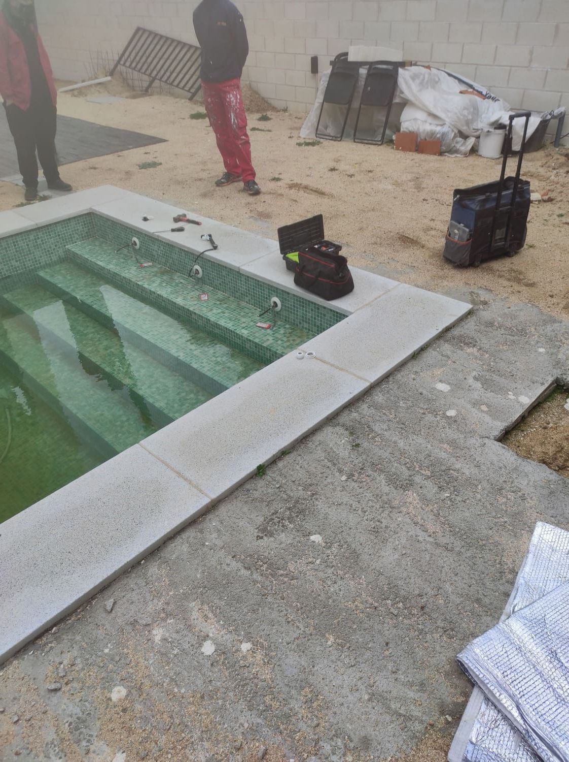 Piscina en construcción con trabajadores cerca, bolsa de herramientas en el borde y escalones que conducen al agua.