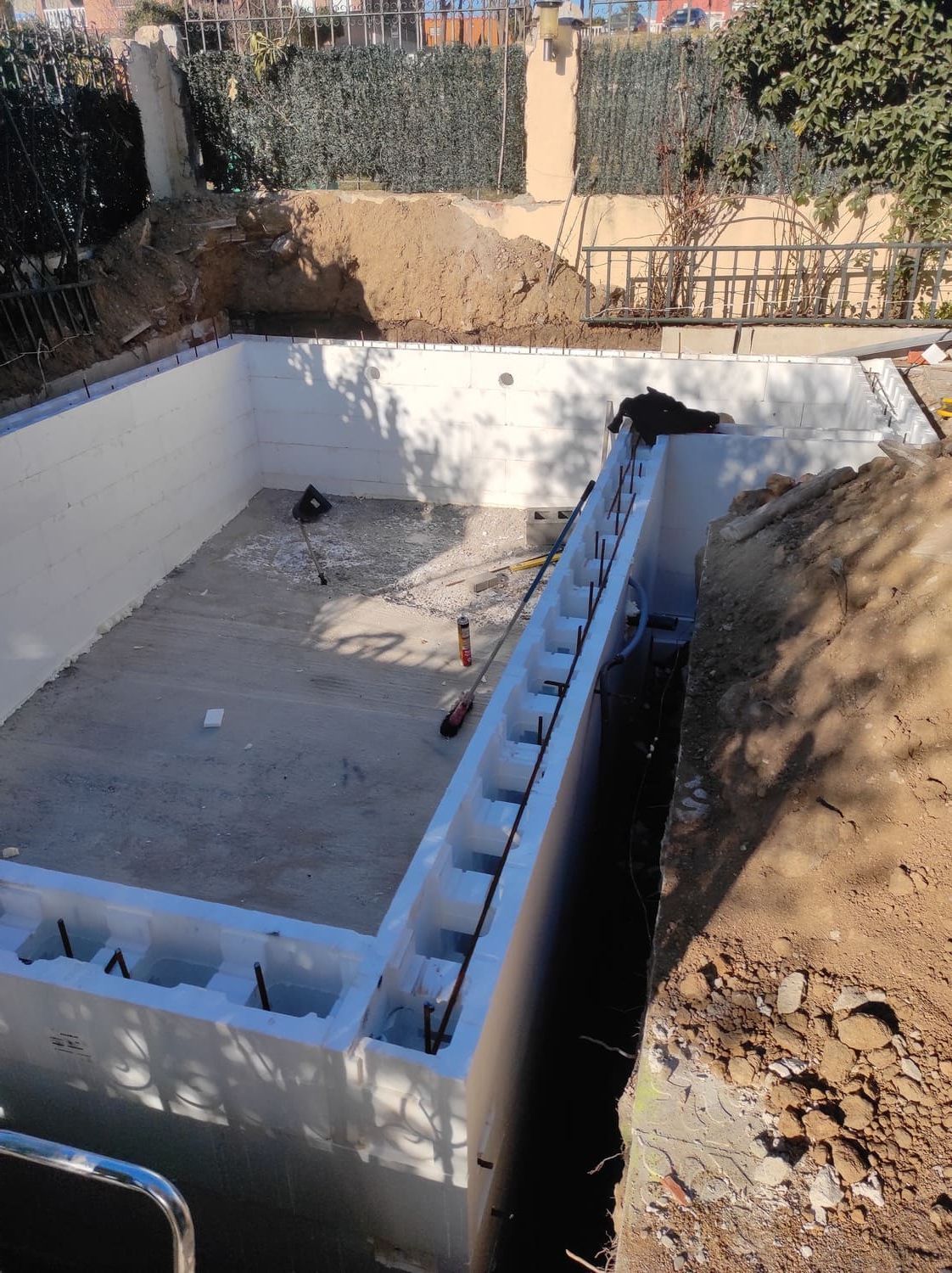 Piscina en construcción; paredes de bloques de espuma blanca, tierra, base de hormigón, entorno exterior soleado.