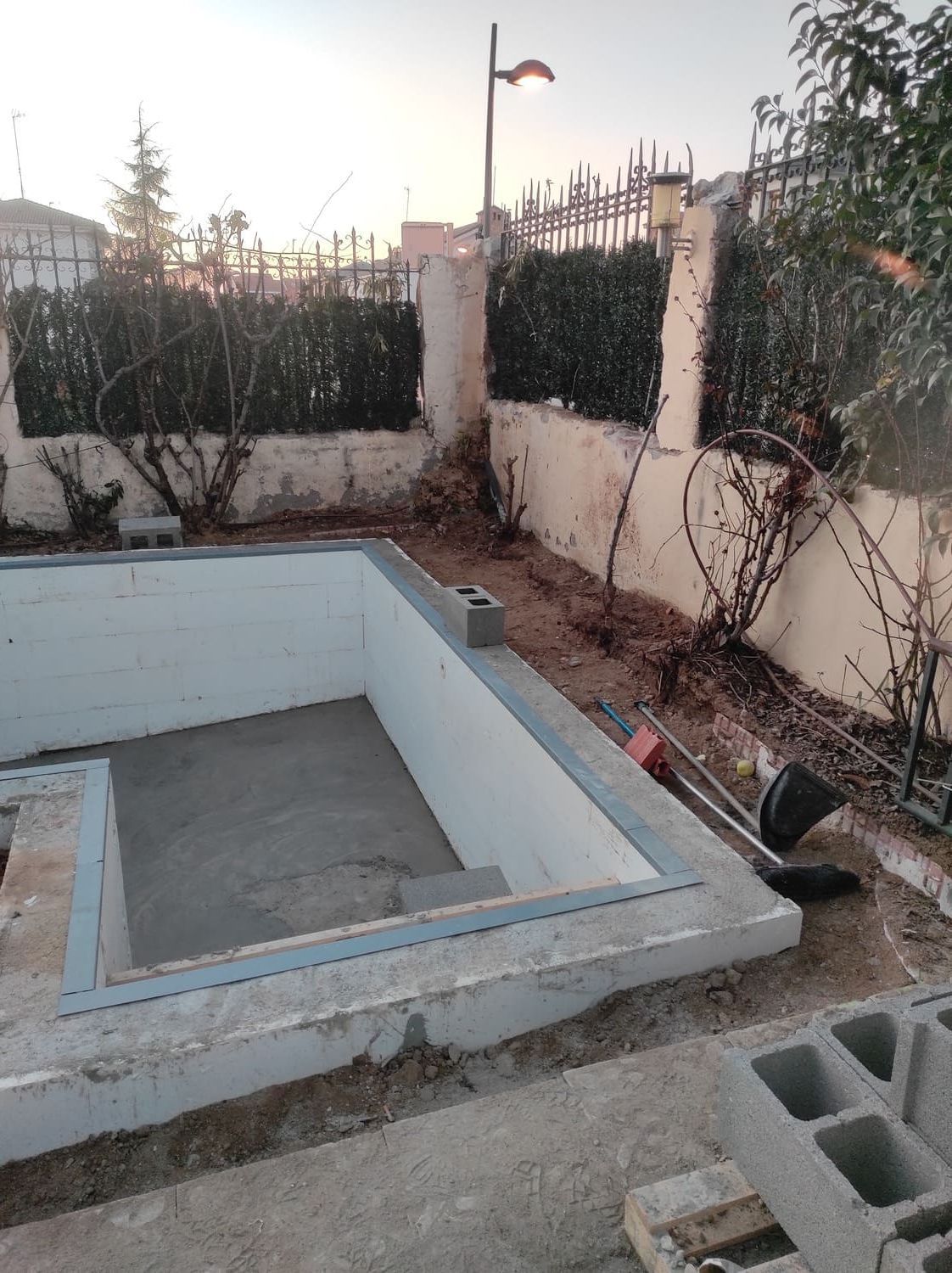 Piscina parcialmente construida. Construcción de cemento gris. Exterior con valla y arbustos.