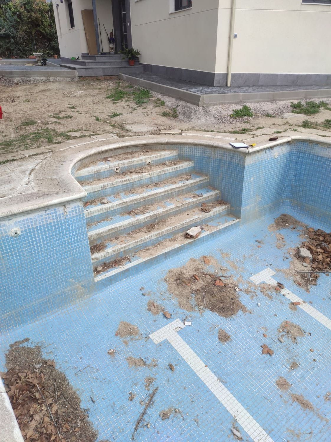 Piscina vacía con azulejos azules y escalones incorporados, suciedad y escombros, al lado de un edificio.