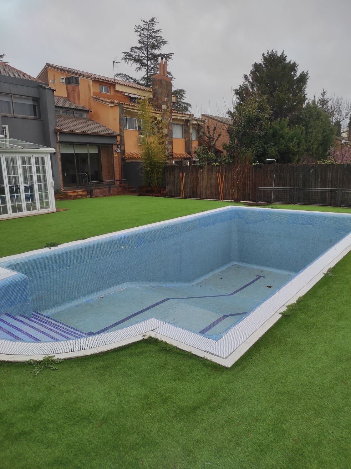 Piscina vacía de azulejos azules rodeada de césped artificial verde, con una casa al fondo en un día nublado.