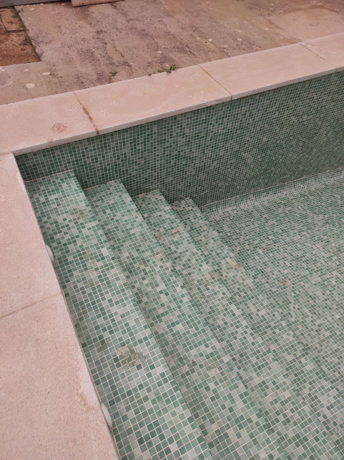Piscina con mosaicos verdes, escalones de hormigón y borde de piedra beige.