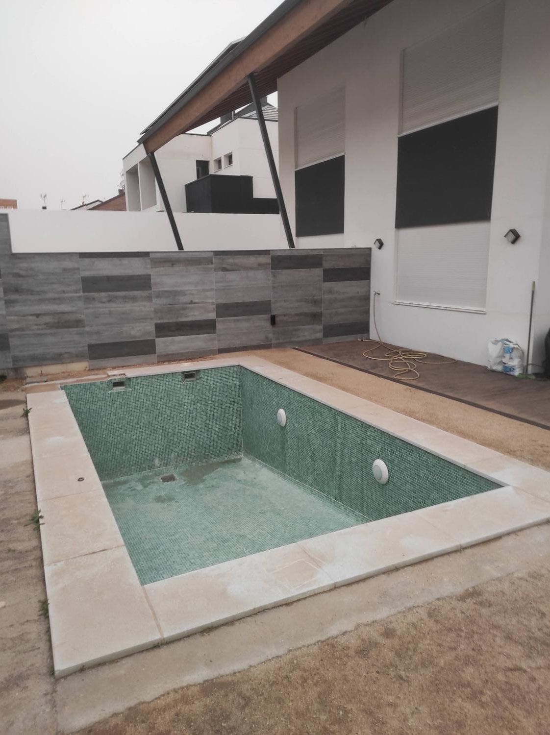 Piscina rectangular vacía con azulejos verdes, al lado de una casa blanca con cortinas oscuras.