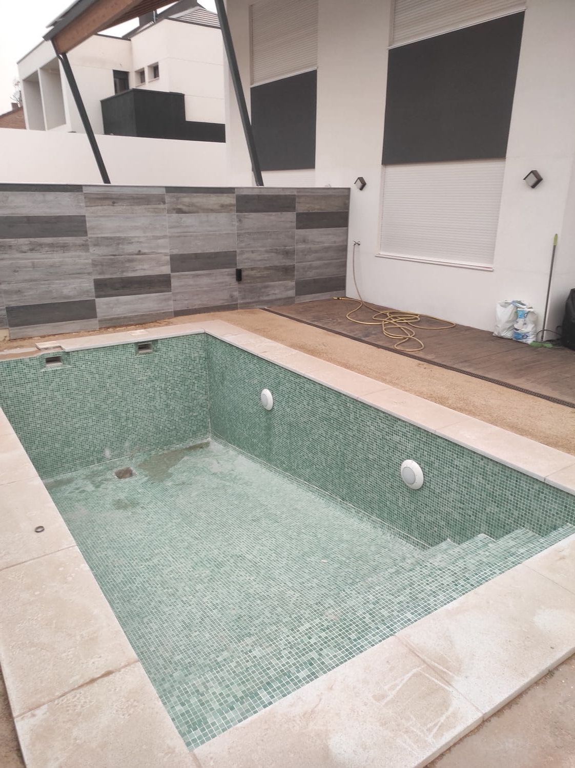 Piscina rectangular con mosaicos verdes, borde beige y escalones, junto a un edificio moderno con detalles en gris y blanco.