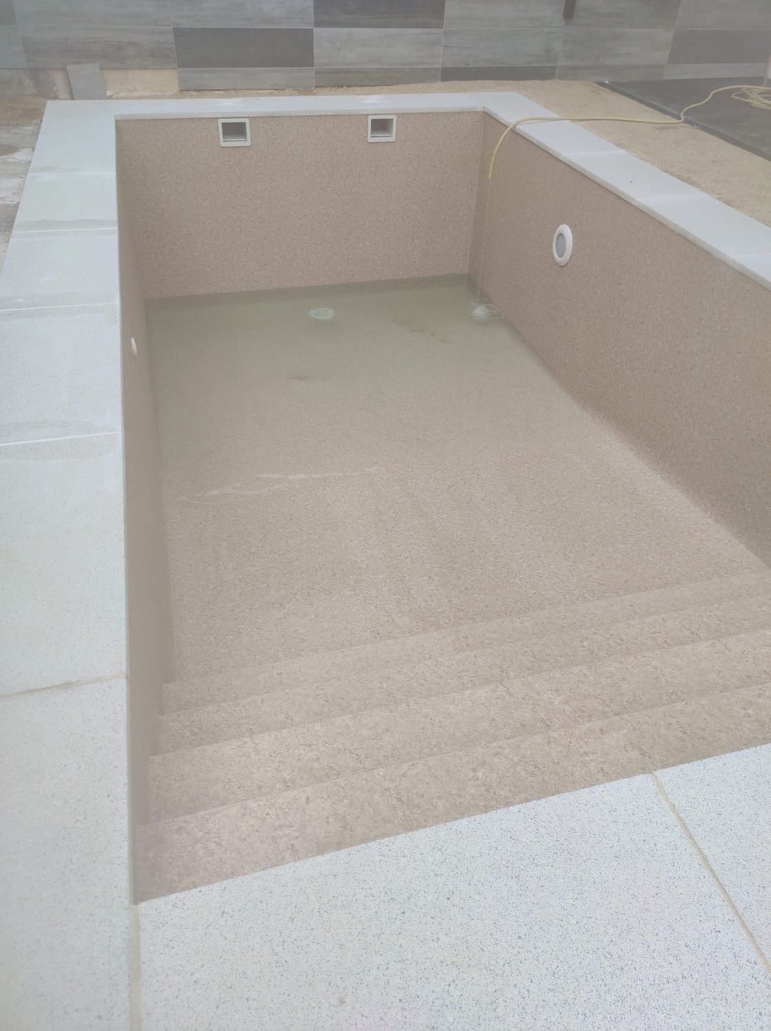 Piscina rectangular de color beige con escalones, rodeada de baldosas de color claro.