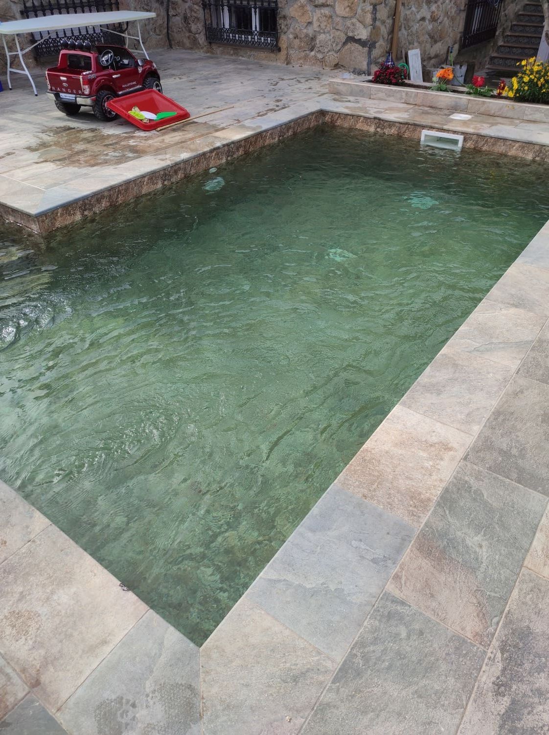 Piscina exterior con revestimiento de piedra y agua verde claro. En el patio hay un coche de juguete rojo y juguetes.