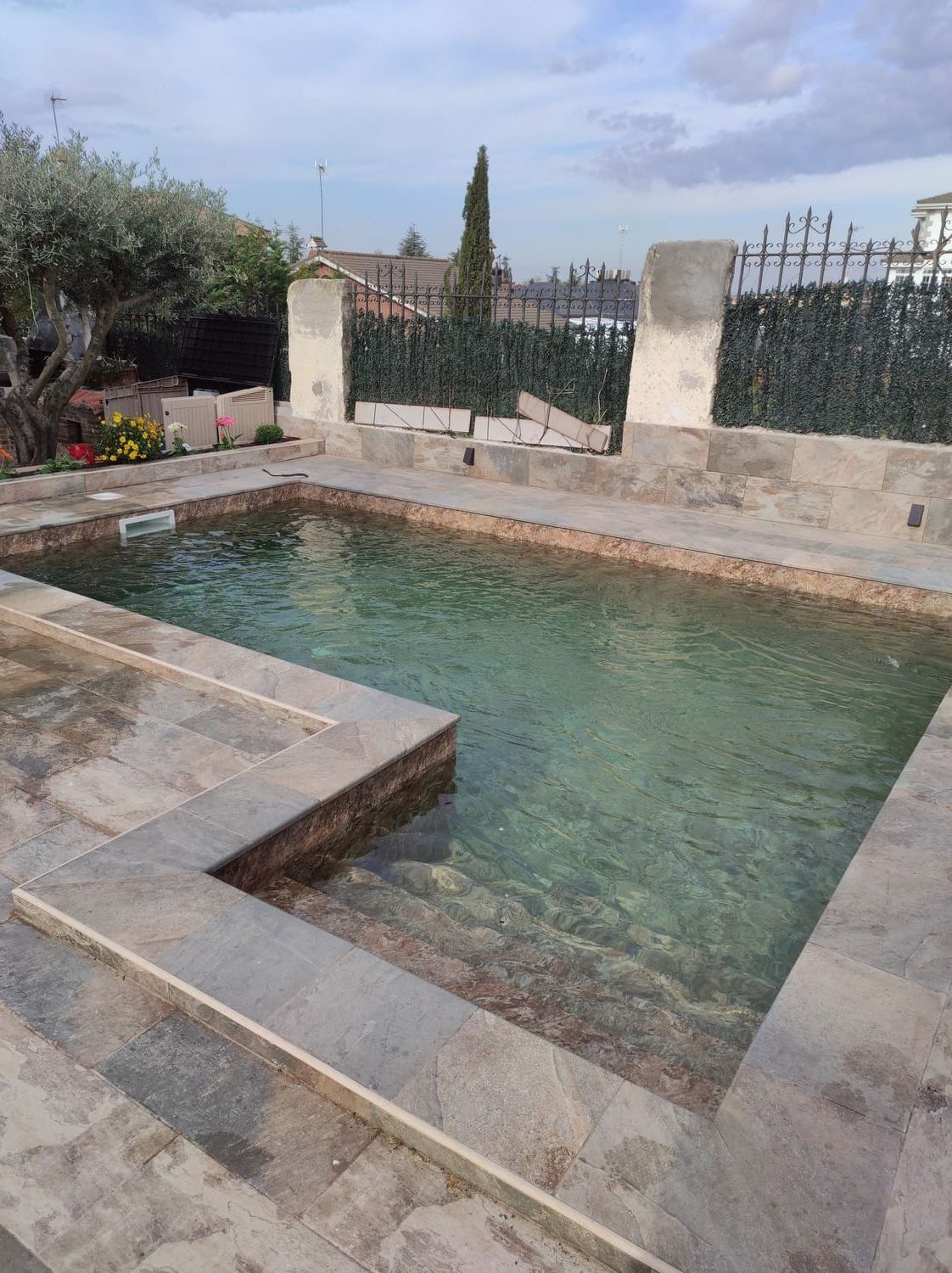 Piscina de piedra con agua cristalina, escalones y patio circundante. Follaje verde y edificios al fondo.